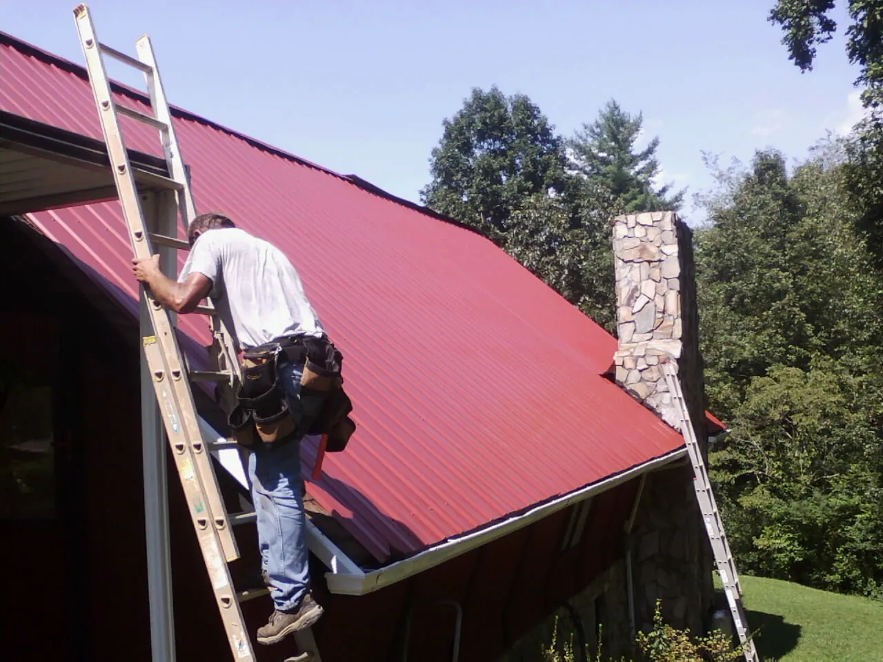 Roofer on a ladder beside a red metal roof and stone chimney — Soffit & Fascia Repair by Roofing Brothers in Kendallville
